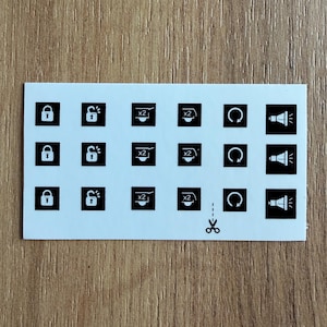 May include: A white sticker sheet with black icons, including padlocks, speaker symbols, and circular arrows. The sheet also features icons with the number "x2" and a scissor symbol for cutting.