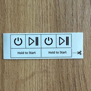 Puede incluir: Una pegatina rectangular blanca con dos conjuntos idénticos de iconos y texto en negro. Cada conjunto incluye un símbolo de encendido, un símbolo de reproducción-pausa y el texto "Hold to Start" dentro de líneas discontinuas.