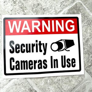May include: A rectangular white sticker with a black border. The top section is red with the word "WARNING" in white. Below, the text "Security Cameras In Use" is displayed with a security camera graphic.