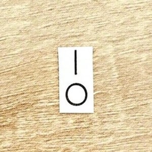 May include: A small, rectangular white sticker with a black line above a black circle. The sticker is placed on a light brown, wood-grain surface. The design resembles an on/off switch.