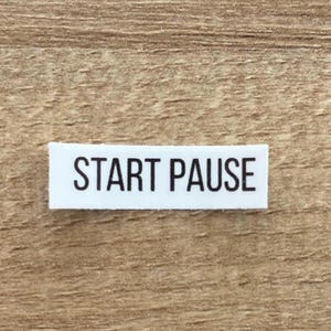 May include: A small, rectangular white label with the words "START PAUSE" printed in black, centered on the label. The label is set against a light brown, wood-grain background.