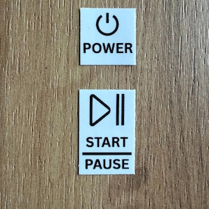 May include: Two white rectangular stickers with black text and symbols on a wood grain surface. The top sticker has a power symbol and the word "POWER". The bottom sticker has a play symbol and the words "START" and "PAUSE".
