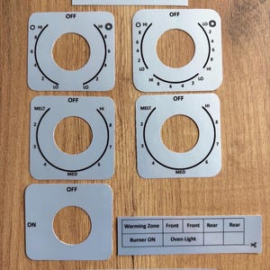 Könnte beinhalten: Ein Set aus hellgrauen Klebeaufklebern für das Bedienfeld eines GE #JB860SJ6SS Herdes. Das Set enthält fünf quadratische Drehknopfumrandungen mit schwarzen Markierungen für Hitzestufen, einen rechteckigen Beschriftungsstreifen und eine Leiste mit Symbolen.
