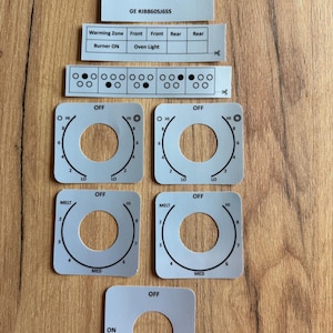 May include: A set of light grey replacement stickers for a GE JB860SJ6SS range control panel. The stickers feature black text and graphics for burner controls, warming zones, and oven light indicators.