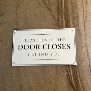 Op de afbeelding: Een rechthoekig wit bord met een dunne zwarte rand en decoratieve hoekaccenten. De tekst in zwarte hoofdletters luidt: PLEASE ENSURE THE DOOR CLOSES BEHIND YOU, gecentreerd tussen twee dunne horizontale lijnen met kleine ruitvormen.