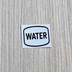 May include: A small, rectangular sticker with rounded corners. The word "WATER" is printed in bold, black letters inside a black, rounded rectangle on a white background. The sticker is on a light wood-grain surface.
