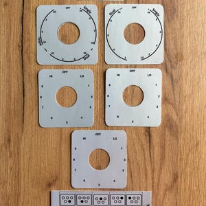 May include: A set of silver-colored stove control panel parts with black markings and text. The parts include circular dials with numbers and labels like "HI," "LO," and "OFF," plus a rectangular strip with burner indicators.
