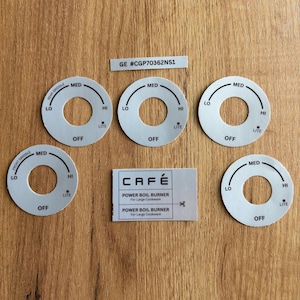 Könnte beinhalten: Ein Set aus fünf weißen, runden Aufklebern mit schwarzer Schrift für Herdknöpfe, inklusive der Markierungen OFF, LITE, LO, MED und HI. Ein Aufkleber ist mit REAR GRIDDLE, einer mit FRONT GRIDDLE und zwei rechteckige Etiketten mit CAFÉ POWER BOIL BURNER beschriftet.