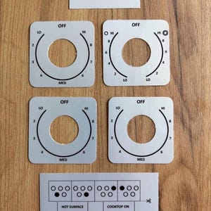 Op de afbeelding: Vier witte vierkante overlays voor fornuisknoppen met zwarte markeringen voor OFF, LO, MED, HI en de cijfers 2 tot 8. Inclusief een rechthoekig label met Samsung #NE59K3310SS/AC en een strook met diagrammen voor indicatielampjes.