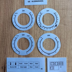 May include: Four white circular stove control knob overlays with black text and markings, including settings like OFF, WARM, HI, LO, and numbers. Includes two rectangular labels with text and diagrams for burner and oven light controls, plus GE #JS968SKSS.