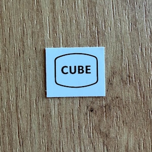 Puede incluir: Una pequeña pegatina cuadrada con fondo azul claro y contorno negro de un rectángulo redondeado. La palabra "CUBE" está impresa en letras negras en negrita dentro de la forma. La pegatina está colocada sobre una superficie de madera.