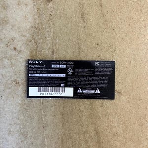 May include: A black rectangular Sony PlayStation 2 product label with white text. It displays the model number SCPH-70012, serial number FS18411113, and a barcode with the number PK218411131. It includes technical specifications, safety warnings, and logos.