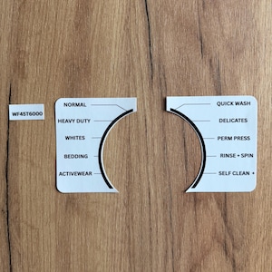 May include: White appliance control panel with the text "NORMAL", "HEAVY DUTY", "WHITES", "BEDDING", and "ACTIVEWEAR". The panel is on a wood grain surface. A separate panel has "QUICK WASH", "DELICATES", "PERM PRESS", "RINSE + SPIN", and "SELF CLEAN +" text.