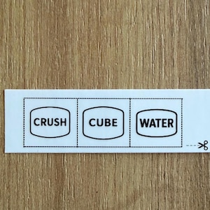 May include: A white label with three sections, each containing a word in a rounded rectangle: "CRUSH", "CUBE", and "WATER". The label has a dotted line for cutting and a scissors icon. The background is a light brown wood grain.