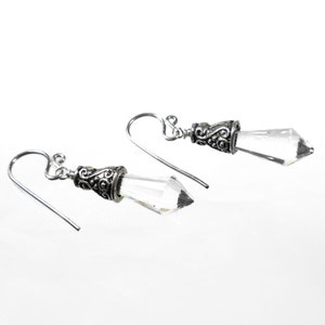 Puede incluir: Un par de pendientes colgantes con cristales transparentes en forma de punta, suspendidos de tapas de metal plateado envejecido con intrincados patrones de volutas y ganchos curvos simples para sujetarlos a la oreja.