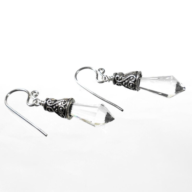 Puede incluir: Un par de pendientes colgantes con cristales transparentes en forma de punta, suspendidos de tapas de metal plateado envejecido con intrincados patrones de volutas y ganchos curvos simples para sujetarlos a la oreja.