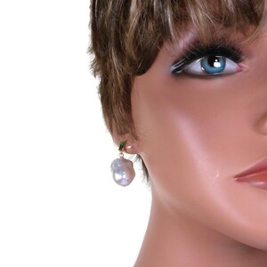 May include: A pair of gold earrings with a single large white baroque pearl dangling from each earring. The earrings are accented with small green gemstones.