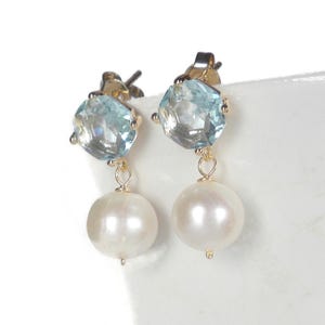 May include: A pair of gold-toned stud earrings. Each earring features a light blue, faceted gemstone above a dangling, round, white pearl. The earrings are set against a white background.