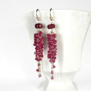 May include: Dangling earrings with a cluster of small, deep red beads suspended from gold-colored hooks. Each earring features a larger, oval-shaped red bead at the top. The earrings are displayed against a white background.