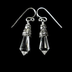 Puede incluir: Un par de pendientes colgantes con cristales transparentes facetados en forma de punta, suspendidos de tapas de metal plateado ornamentadas con volutas intrincadas y ganchos de alambre para orejas perforadas.