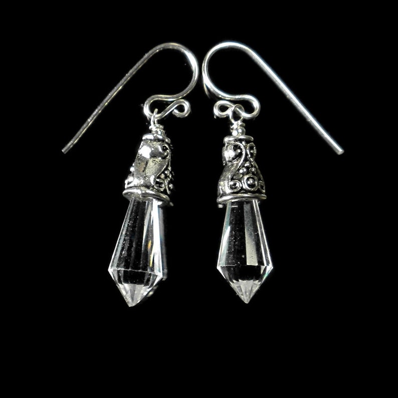 Puede incluir: Un par de pendientes colgantes con cristales transparentes facetados en forma de punta, suspendidos de tapas de metal plateado ornamentadas con volutas intrincadas y ganchos de alambre para orejas perforadas.