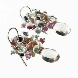 May include: A pair of handcrafted earrings featuring clusters of white pearls and colorful gemstones, including ruby, sapphire, and emerald. Each earring has a large, clear, teardrop-shaped crystal pendant and gold-colored wire accents.
