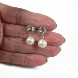 May include: A pair of gold-toned earrings featuring a light blue, faceted gemstone at the top and a large, round, cream-colored pearl dangling below. The earrings are displayed against a white background.