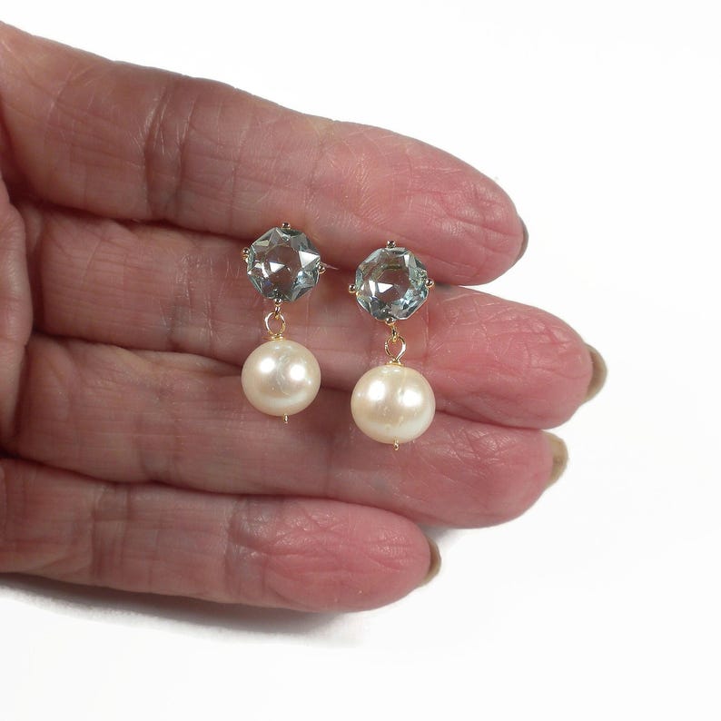 May include: A pair of gold-toned earrings featuring a light blue, faceted gemstone at the top and a large, round, cream-colored pearl dangling below. The earrings are displayed against a white background.