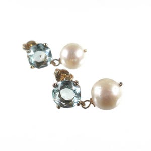 May include: A pair of gold-toned earrings, each featuring a light blue, square-cut gemstone and a large, round, white pearl. The earrings are displayed against a white background, highlighting the jewelry's details.