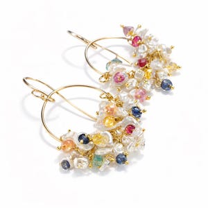 May include: A pair of gold hoop earrings featuring clusters of white irregular freshwater pearls and small faceted gemstones in shades of blue, yellow, pink, and red. The stones are attached to the thin gold wire hoops with delicate gold links.
