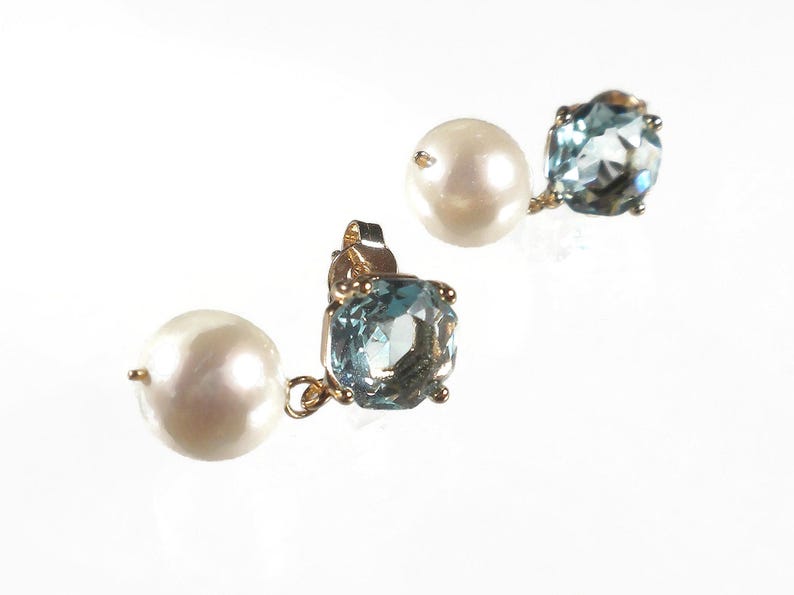 May include: A pair of gold-toned stud earrings. Each earring features a large, round, white pearl and a light blue, square-cut gemstone. The gemstones have multiple facets. The earrings are set against a white background.