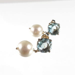 May include: A pair of gold earrings featuring a large, round, white pearl and a square, light blue gemstone. The earrings are set on a gold backing and are displayed against a white background.