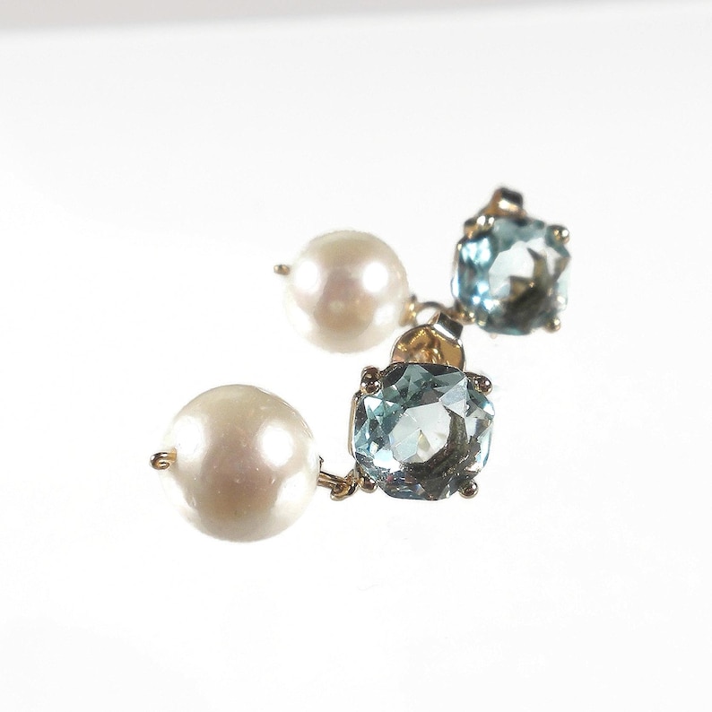 May include: A pair of gold earrings featuring a large, round, white pearl and a square, light blue gemstone. The earrings are set on a gold backing and are displayed against a white background.