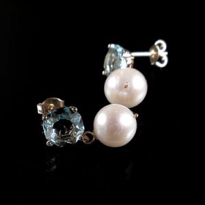 May include: A pair of stud earrings with a light blue gemstone and a white pearl. The earrings are set in gold and are displayed against a black background. The gemstones are a light blue color.