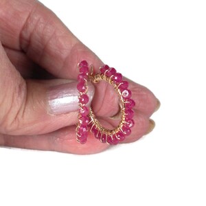 May include: A small hoop earring constructed from thin gold-toned wire wrapped with numerous tiny, faceted, vibrant magenta-pink beads. The beads are densely arranged along the entire circumference of the circular wire frame.