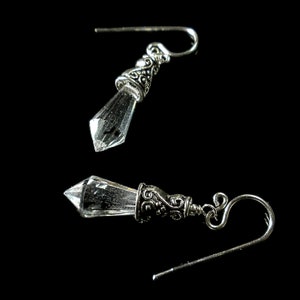 Puede incluir: Un par de pendientes colgantes con cristales transparentes facetados en forma de punta, suspendidos de tapas de metal plateado adornadas con intrincados patrones de volutas y ganchos curvos para la oreja.