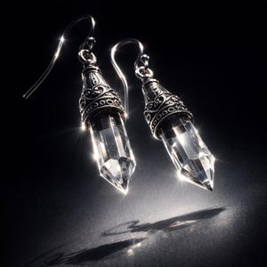 Puede incluir: Un par de pendientes colgantes con cristales transparentes tallados en punta, suspendidos de tapas met&aacute;licas plateadas ornamentadas con intrincados patrones de volutas y ganchos curvos para las orejas.