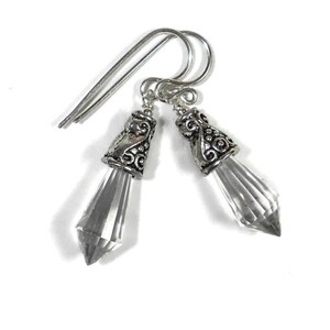 Puede incluir: Un par de pendientes colgantes con cristales transparentes facetados en forma de punta, suspendidos de casquillos de metal plateado con intrincados grabados de volutas y c&iacute;rculos, unidos a ganchos de metal plateado para orejas perforadas.