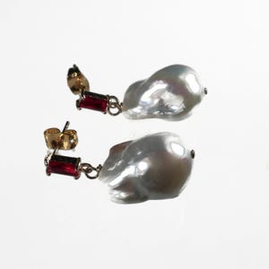 May include: A pair of earrings featuring a large white baroque pearl with a red gemstone dangle. The earrings have gold-tone posts and are a statement piece.