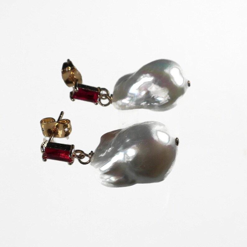 May include: A pair of earrings featuring a large white baroque pearl with a red gemstone dangle. The earrings have gold-tone posts and are a statement piece.