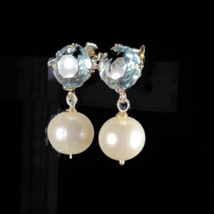 May include: A pair of elegant earrings featuring light blue, faceted gemstones and large, round, cream-colored pearls. The earrings are suspended from gold-colored metal settings, creating a classic and sophisticated look. The background is black.