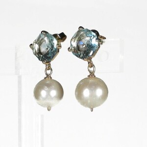 May include: A pair of gold-toned earrings. Each earring features a light blue, square-cut gemstone at the top, connected to a large, round, white pearl. The earrings are displayed against a white background.