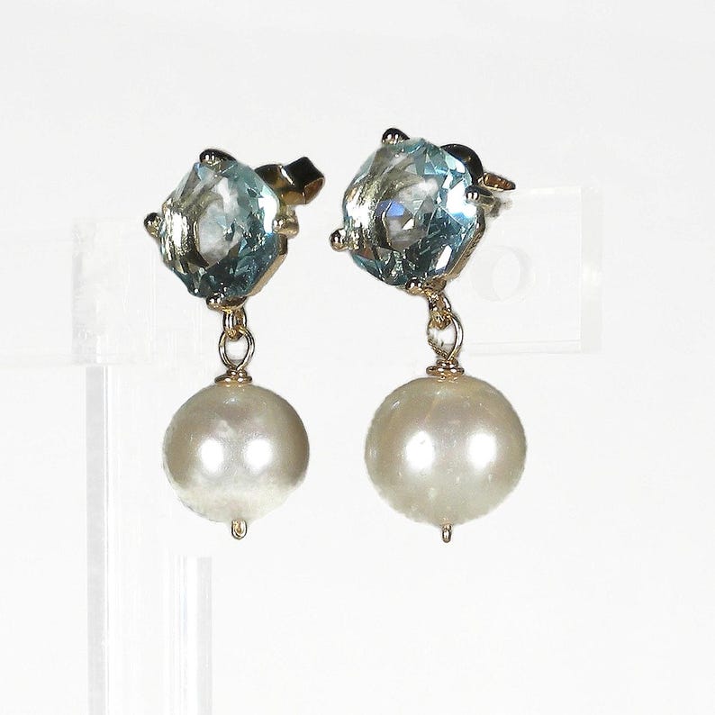 May include: A pair of gold-toned earrings. Each earring features a light blue, square-cut gemstone at the top, connected to a large, round, white pearl. The earrings are displayed against a white background.