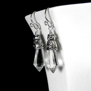 Puede incluir: Un par de pendientes colgantes con gotas de cristal transparentes, facetadas y puntiagudas, suspendidas de tapas de metal plateado ornamentadas con intrincados patrones de volutas y ganchos curvos simples para la oreja.