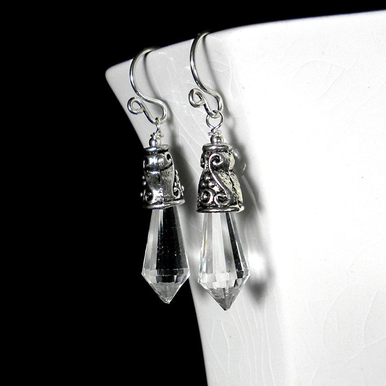 Puede incluir: Un par de pendientes colgantes con gotas de cristal transparentes, facetadas y puntiagudas, suspendidas de tapas de metal plateado ornamentadas con intrincados patrones de volutas y ganchos curvos simples para la oreja.