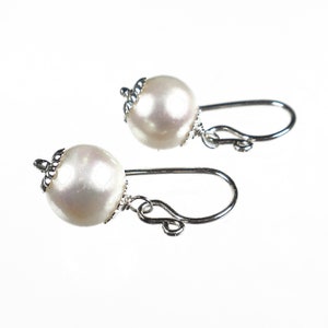 Puede incluir: Un par de pendientes de perlas con ganchos plateados. Cada pendiente presenta una perla blanca redonda y brillante, acentuada con una tapa plateada decorativa. Los ganchos son curvos y están diseñados para orejas perforadas.
