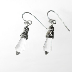 Puede incluir: Un par de pendientes colgantes con cristales transparentes facetados en forma de punta, suspendidos de tapas de metal plateado envejecido con adornos. Los pendientes se sujetan mediante ganchos de metal plateado para orejas perforadas.