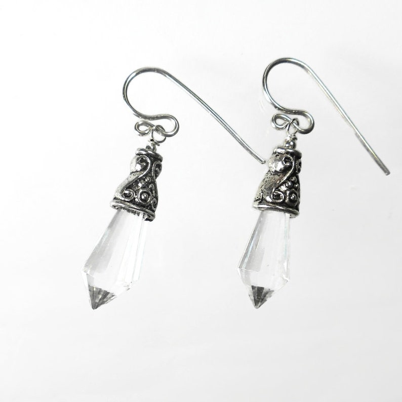 Puede incluir: Un par de pendientes colgantes con cristales transparentes facetados en forma de punta, suspendidos de tapas de metal plateado envejecido con adornos. Los pendientes se sujetan mediante ganchos de metal plateado para orejas perforadas.