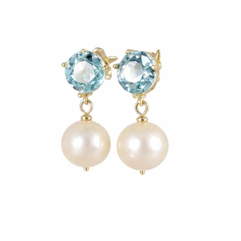May include: A pair of gold earrings featuring a light blue gemstone at the top and a large, round, white pearl dangling below. The earrings are set against a white background.