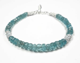 May include: A bracelet featuring faceted blue-green gemstone beads arranged in a circular pattern, interspersed with small, textured silver-toned metal spacer beads. The piece includes a silver-toned metal clasp mechanism at the top for closure.